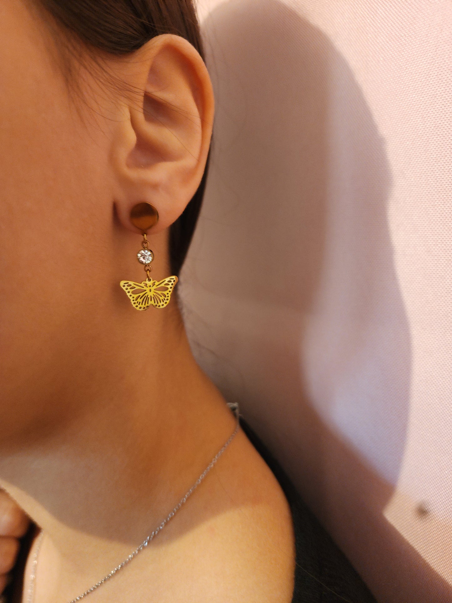 Boucles d'Oreilles Paméla en acier inoxydable doré avec strass brillant et pendentif papillon ajouré, portées sur une oreille